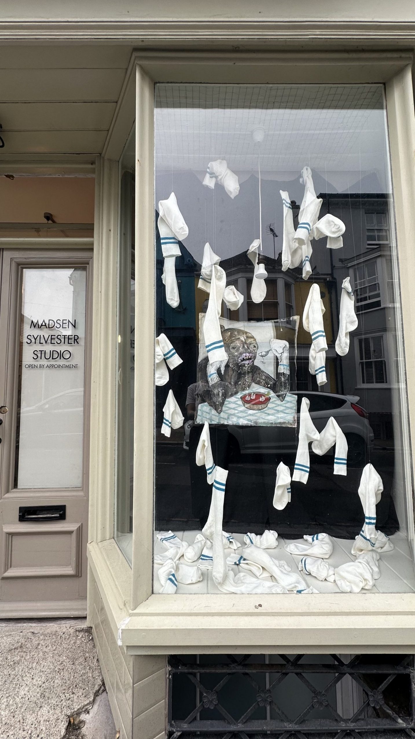 A window display featuring an installation. (Centre) A painted figure with exaggerated features is positioned behind a table with a bowl, the figure is grey and red. Surrounded by suspended white socks with blue stripes. Dozens of similar socks are scattered across the floor of the display.
