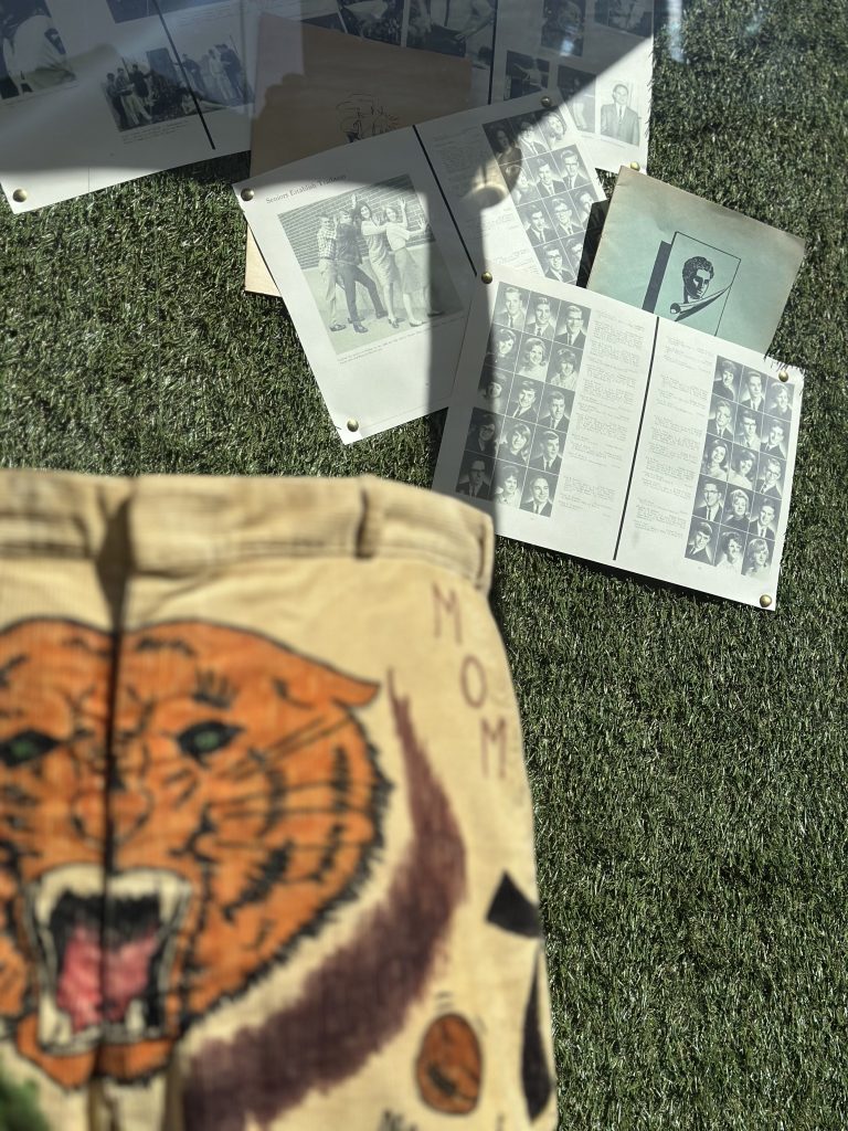 On a flat lay display which is green grass, is a pair of cord yellow trousers with a roaring yellow and black striped tiger on the front. Above the trousers are a series of black and white photographs.