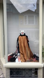 Window display of a long bronze satin cloak over a black and grey layered ensemble, with red embroidery details visible on one sleeve. Various costume pieces, including fabric and accessories, are scattered at the base.