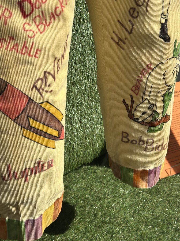 This image is a close up of yellow cord trousers that has a collection of words on each leg, a pink, yellow and purple rocket and a lamb holding a brown branch.