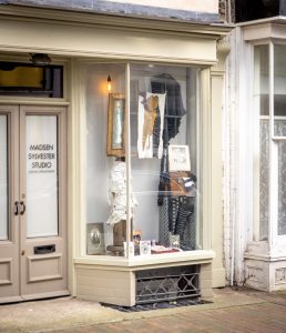 A window display featuring an installation of a collection of objects. At the bottom right hand corner there is a long black boot with laces, and vintage photographs. There is a metal case which can be seen supporting a brown box with a framed vintage photogram in black ad white on top. On the left hand side is a tall wooden block which has layers of white material wrapped around it. Suspended above is a large gold photo frame with a colourful painting and to the right is a black cape. There are paintings stuck to the widow itself.