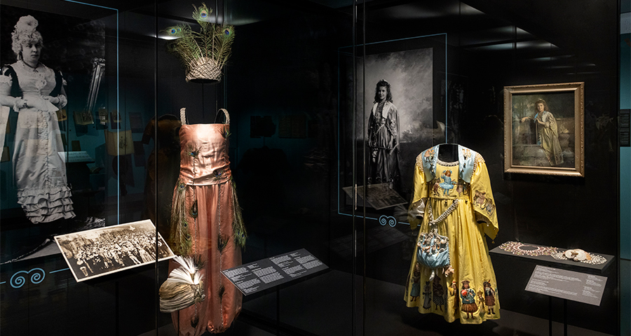(Left) A peach-pink dress adorned with peacock feathers, positioned beneath a feathered headpiece and beside a fan and vintage photograph. (Right) A bright yellow gown decorated with painted scenes and paired with a matching handbag; nearby, a painting and black-and-white portrait show the garment in use.