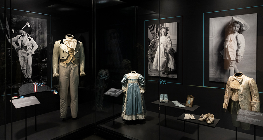 A child’s cream suit with gold a sash and a blue and white child’s dress with puffed sleeves.