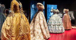 Four female ballgowns displayed on Mannequins. (Left) A yellow dress with silver embellishments and ruffled sleeves. (Centre Left) White with pink floral pattern, and ruffled half sleeves. (Centre Right) Light powder blue dress with embellishments and ruffles throughout. (Right) Pink gown with with white quarter sleeves.