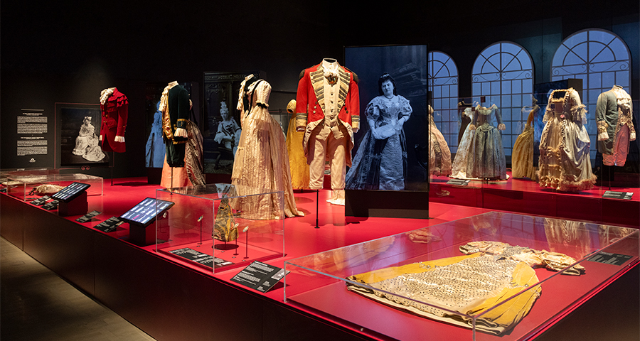 (Left) A male mannequin in a red military jacket with gold details stands beside elaborately dressed female mannequins in embroidered silk gowns. (Centre) A prominent male mannequin wears a red and white uniform-style coat with gold epaulettes, surrounded by gowns in pale pink, gold, and blue tones. (Right) Additional mannequins feature both male and female costumes, including richly decorated bodices, lace-trimmed skirts, and brocade jackets.
