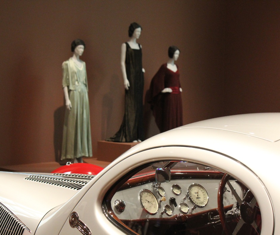 The mannequins are dressed in long, elegant evening dresses from the early 20th century. The dresses are made of soft fabrics in muted green, black, and deep red tones.