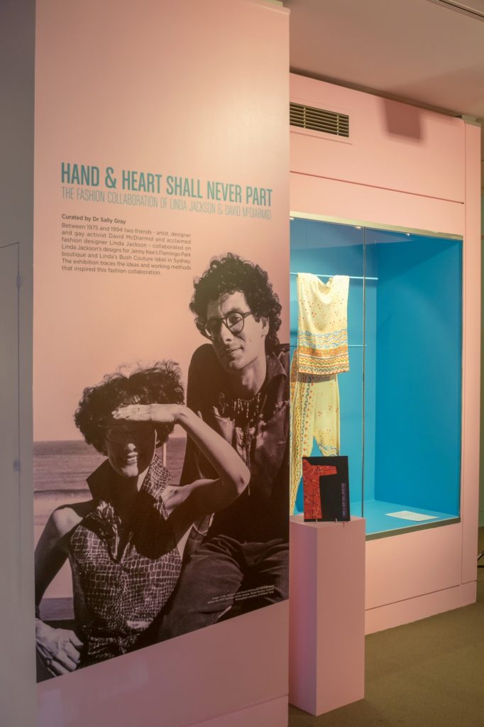 gallery view of colourful walls and garments mounted on walls. Intro text panel with image of curators.
