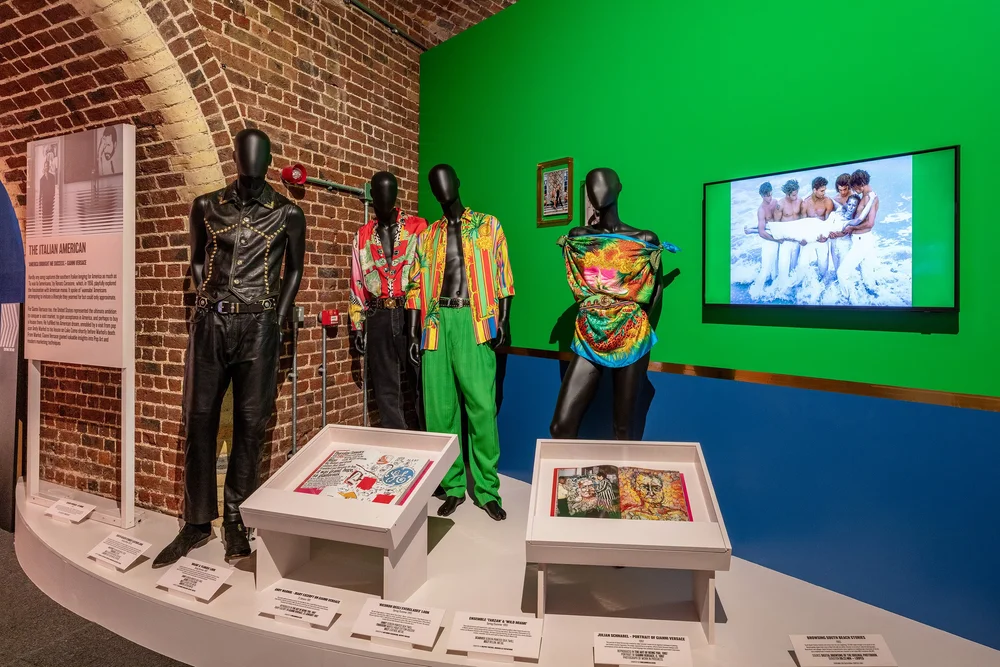 Exhibition display of mannequins in colourful clothing against a green backdrop. Some display cases to front