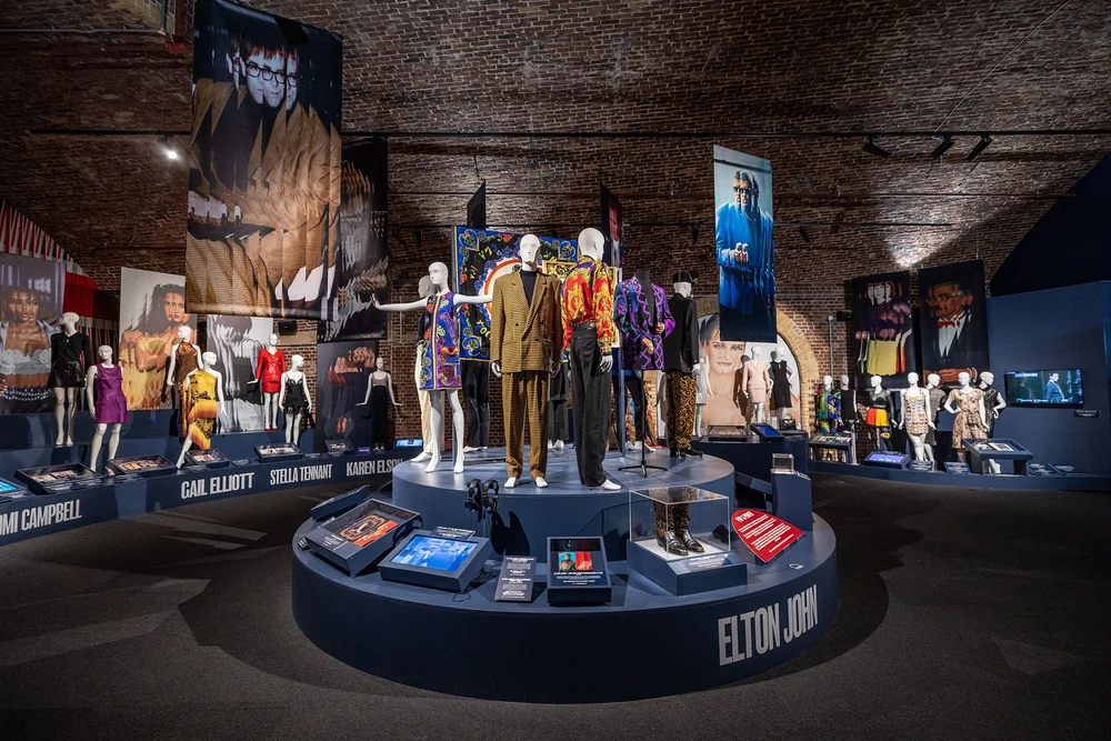 mannequins on a round plinth in middle of brick-walled gallery with more mannequins in the background, each plinth named with a celebrity wearer.