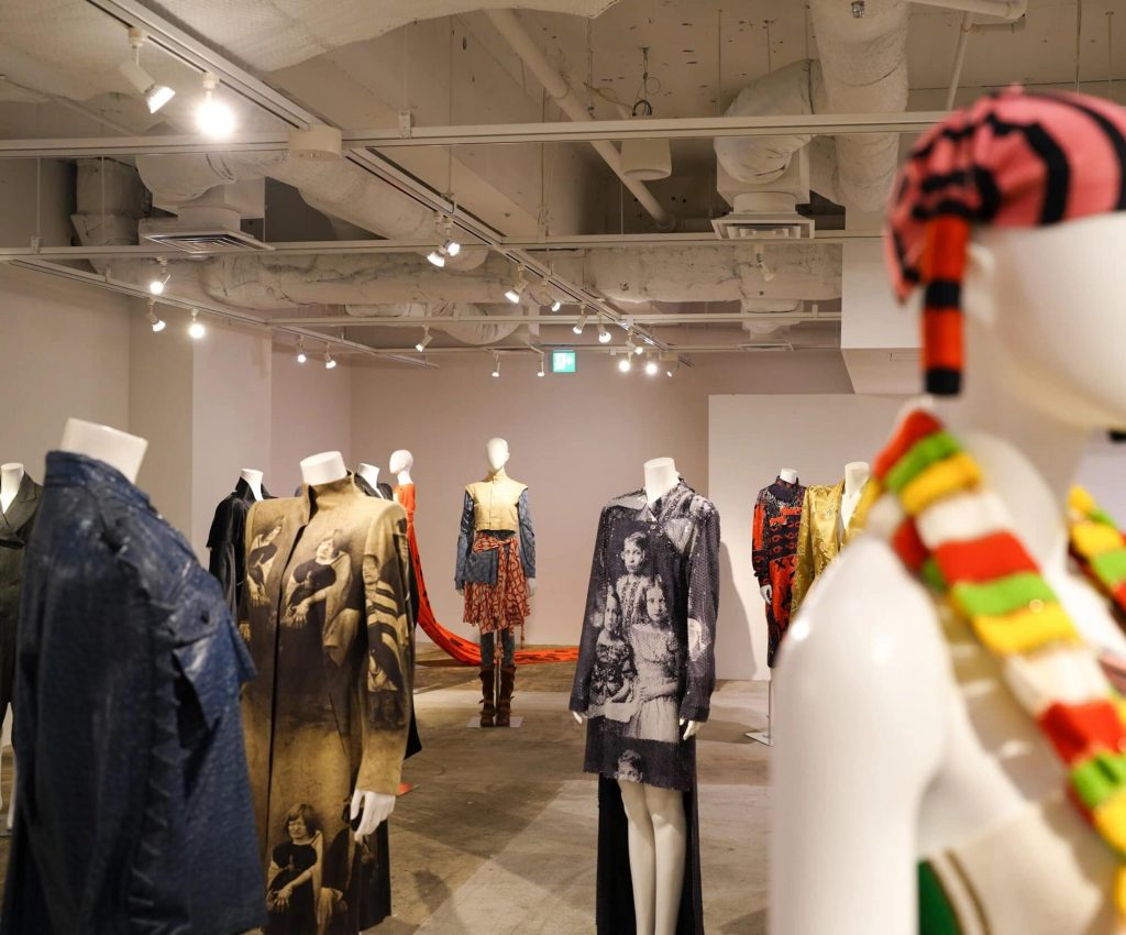 The clothes have some featuring printed photos of people. One mannequin wears a colourful scarf and hat, while others have outfits with bold shapes and patterns.