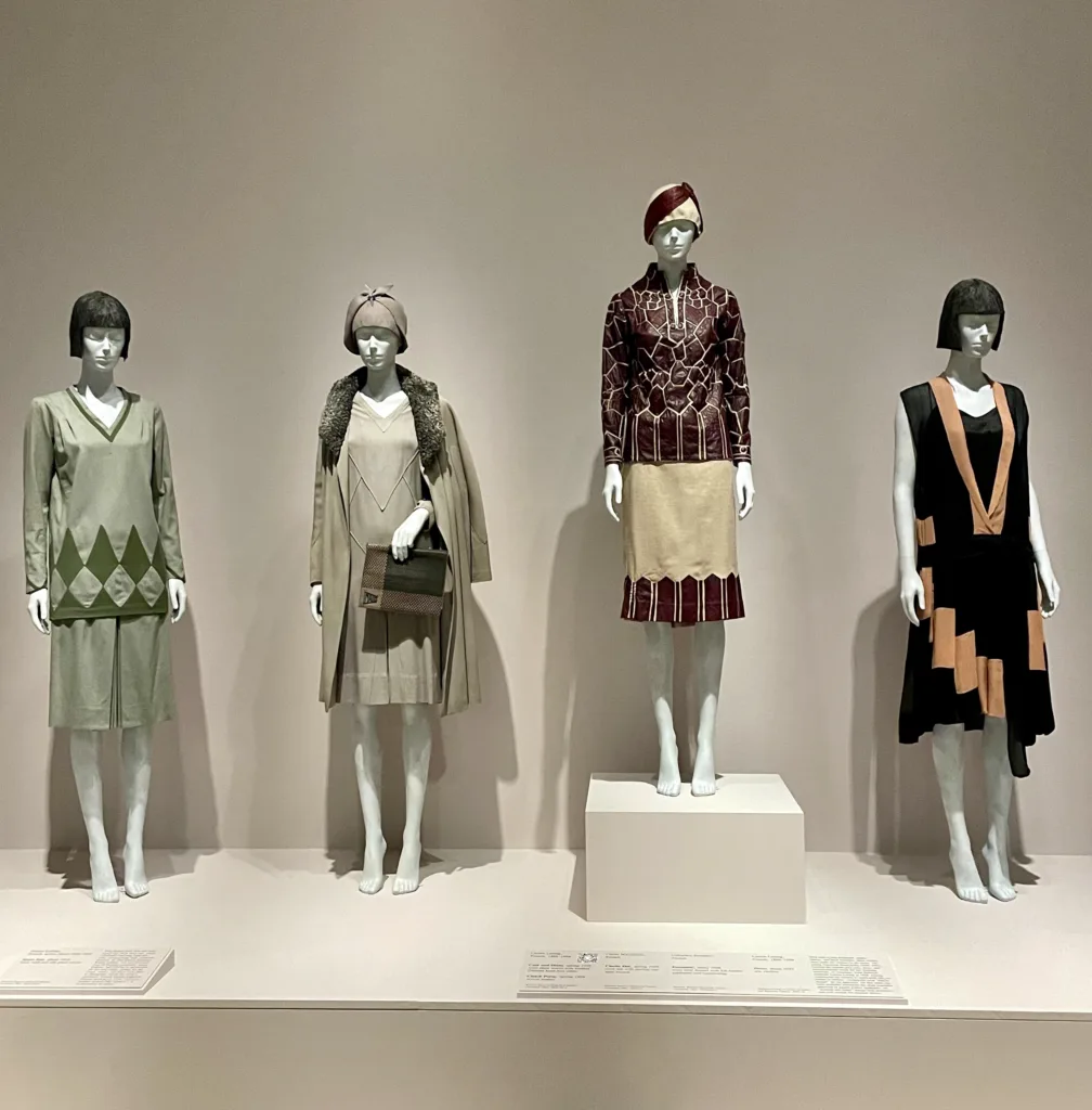 The mannequins are dressed in 1920s-style outfits with simple shapes and bold geometric patterns. The dresses use muted and rich colours, and some include matching hats or capes.