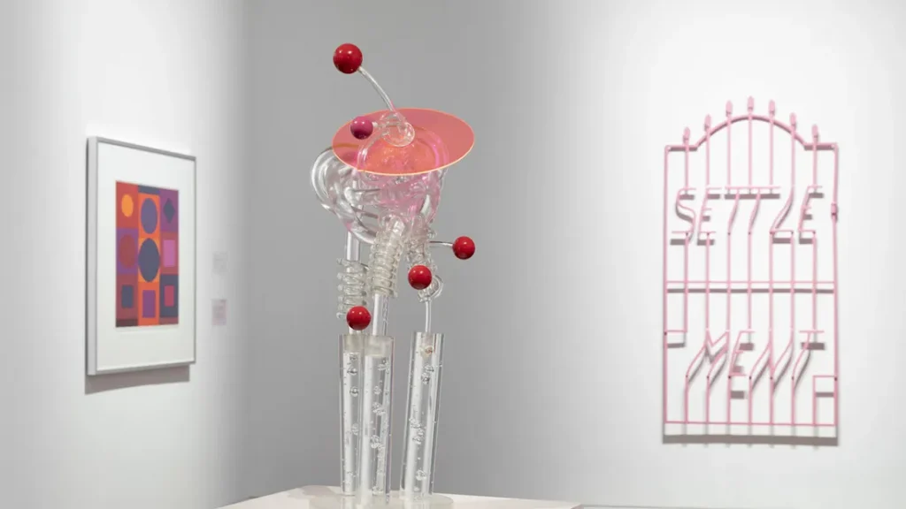 gallery display of sculptural object with a framed abstract painting and a metal walll hanging with the word settlement in background