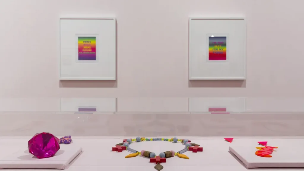 gallery vitrine display of jewellery and objects with framed pictures of slogans against a rainbow colour