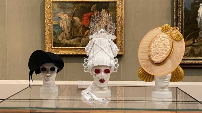 Three hats on display in hues of black, white and yellow. One of the mannequins wear round red glasses.